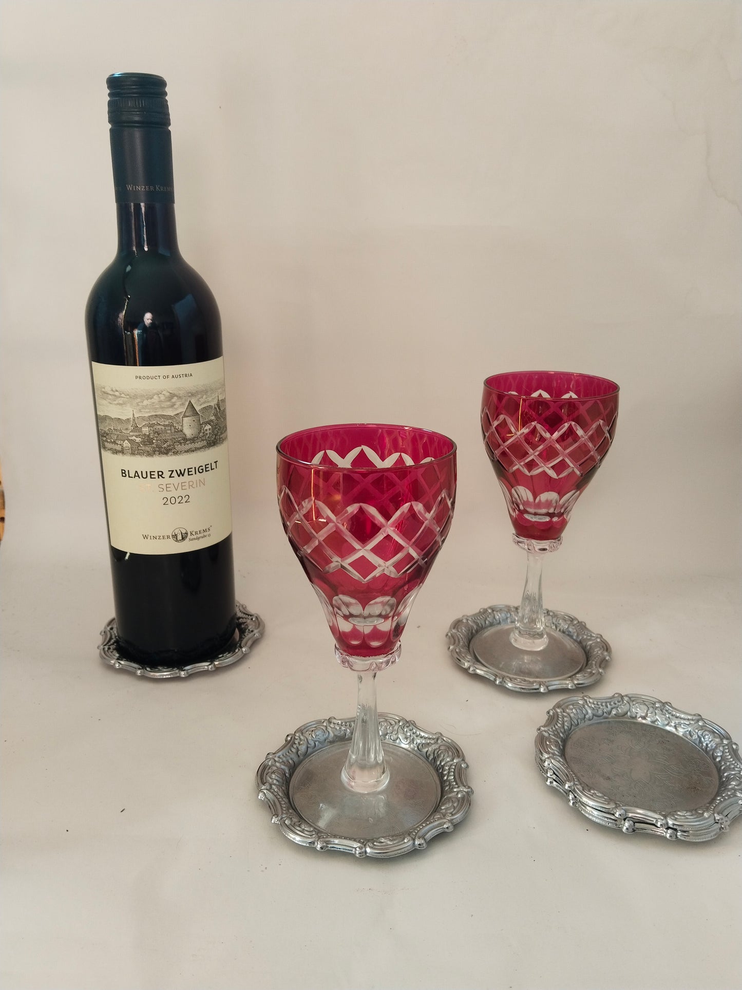 Anwendungsbeispiel: Eine Flasche Wein und zwei rubinrote Kristallgläser auf den versilberten Untersetzern.