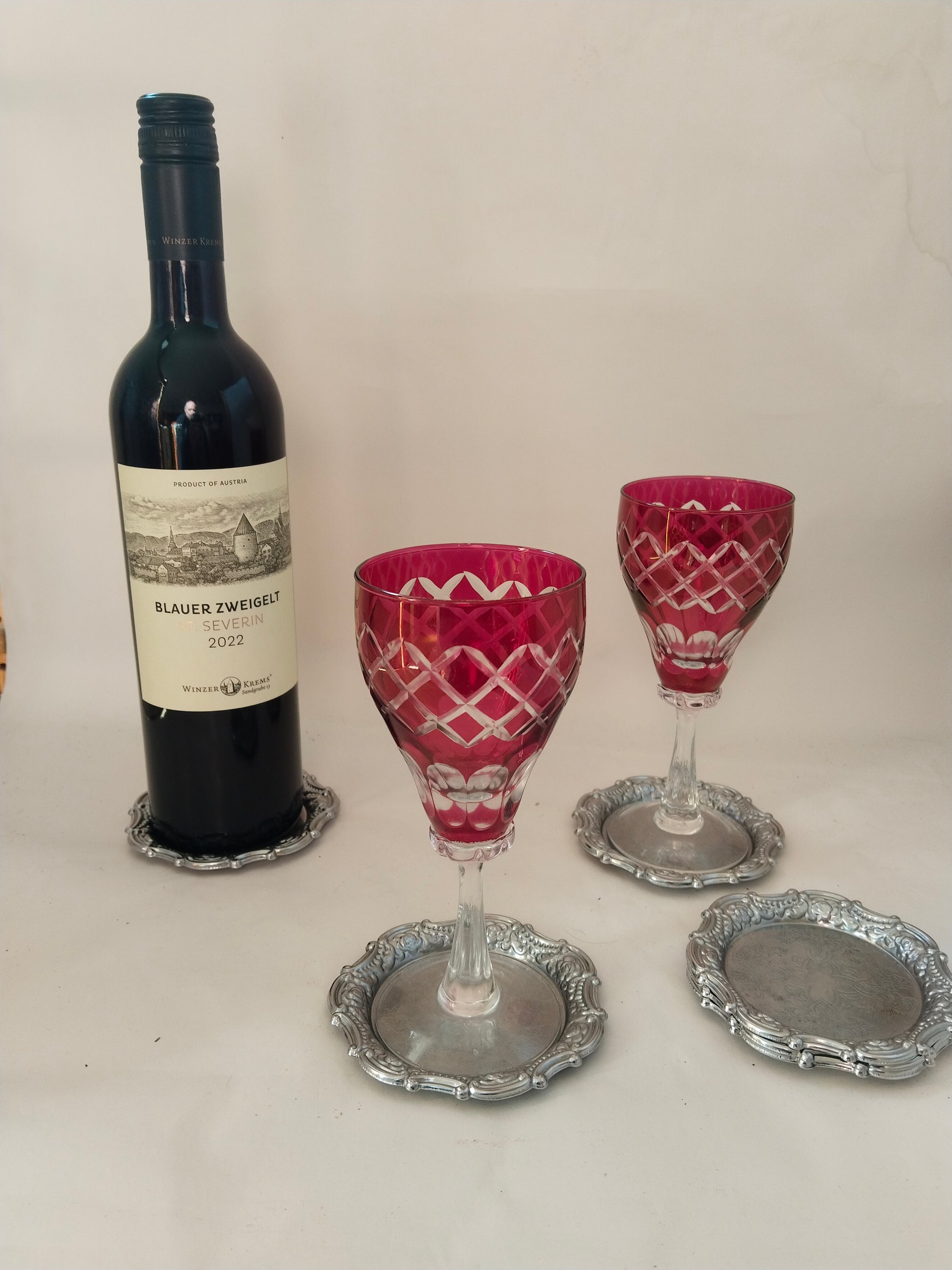 Anwendungsbeispiel: Eine Flasche Wein und zwei rubinrote Kristallgläser auf den versilberten Untersetzern.