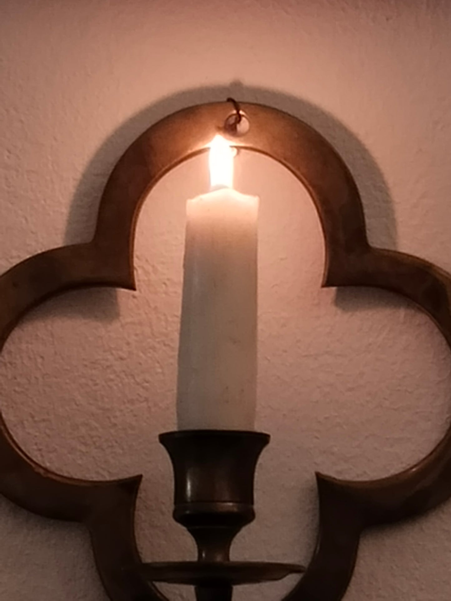 Historischer Wandleuchter aus Messing im Vierpass-Design an einer weißen Wand mit brennender Kerze.