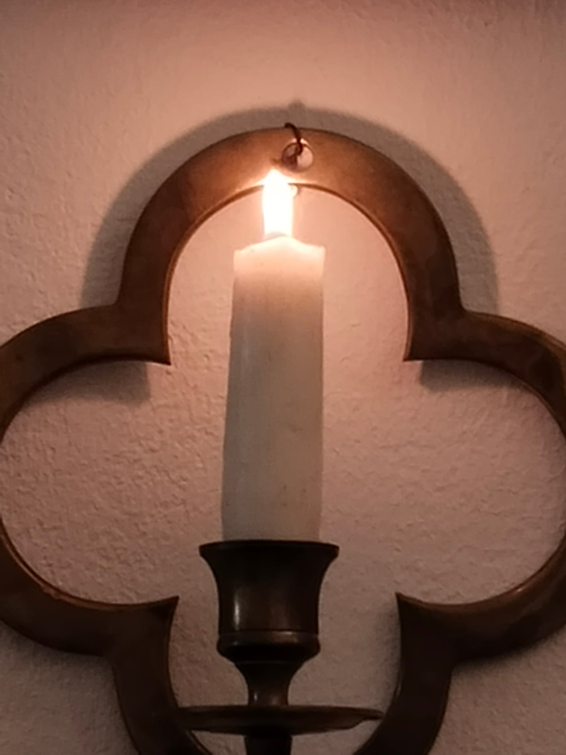 Historischer Wandleuchter aus Messing im Vierpass-Design an einer weißen Wand mit brennender Kerze.