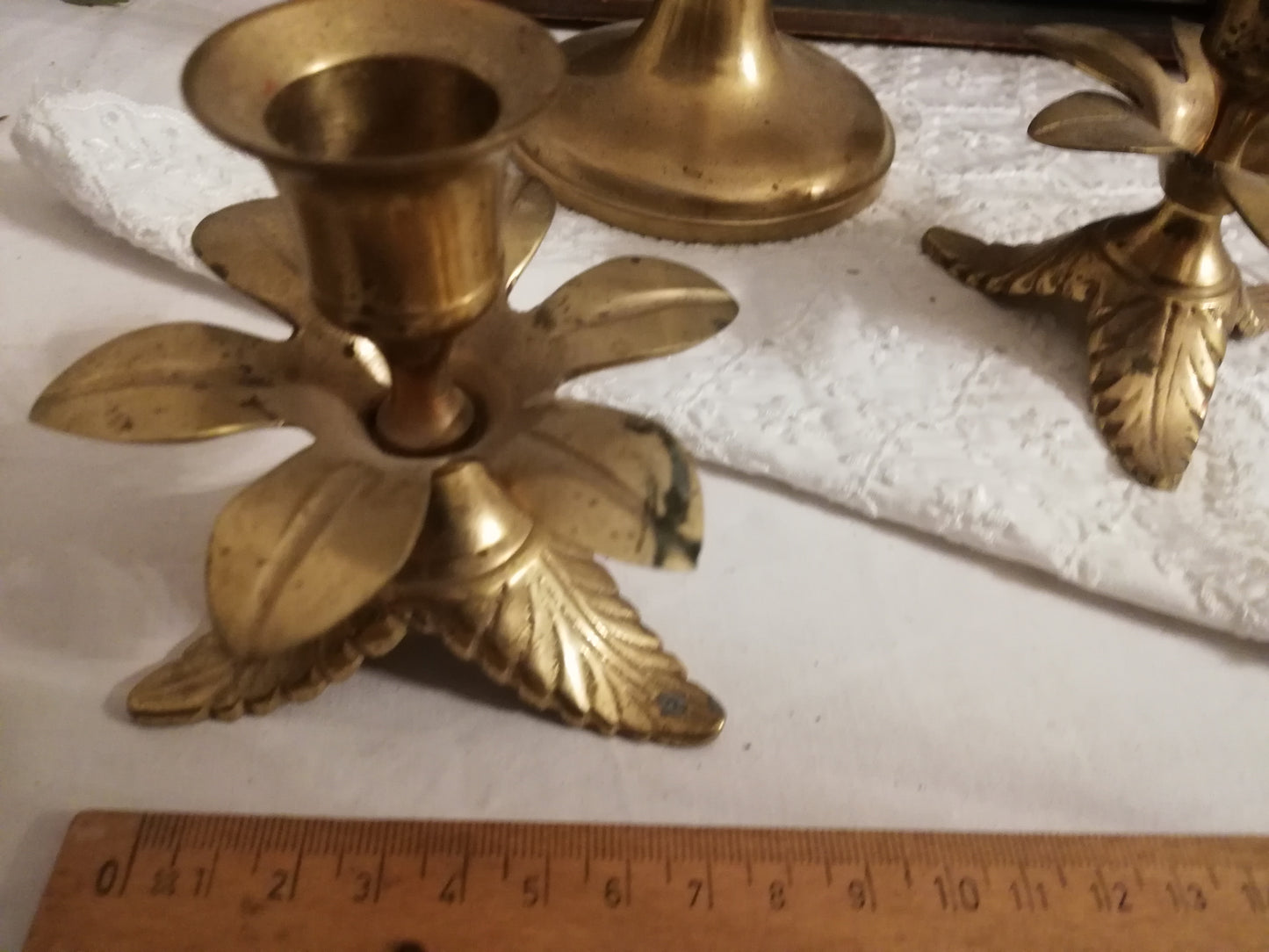 Zwei antike Messing-Kerzenständer mit Akanthusblatt-Fuß auf weißer Spitzendecke. zusammen mit einem Lineal.