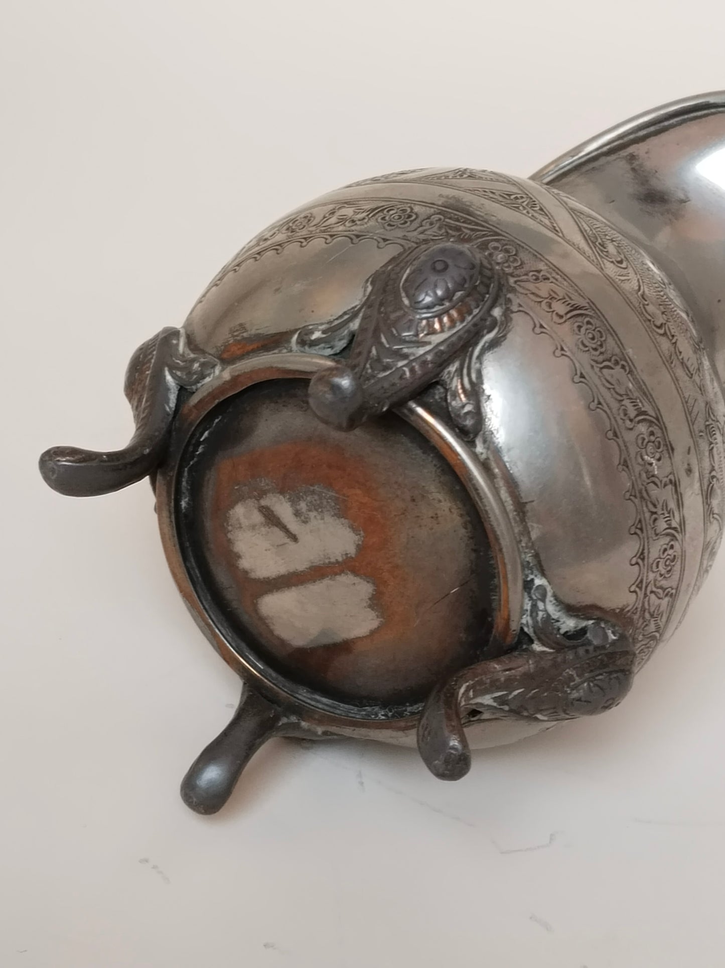 Unterseite des Milchkännchens mit Standfuß, abgenutzte Stelle mit kupferfarben durchschimmerndem Material
