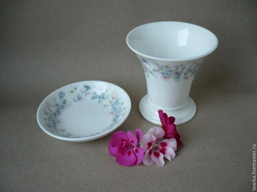 Frontansicht einer weißen Wedgwood Vase und eines kleinen Tellers mit floralem Angela-Dekor und pinken Blüten im Vordergrund.
