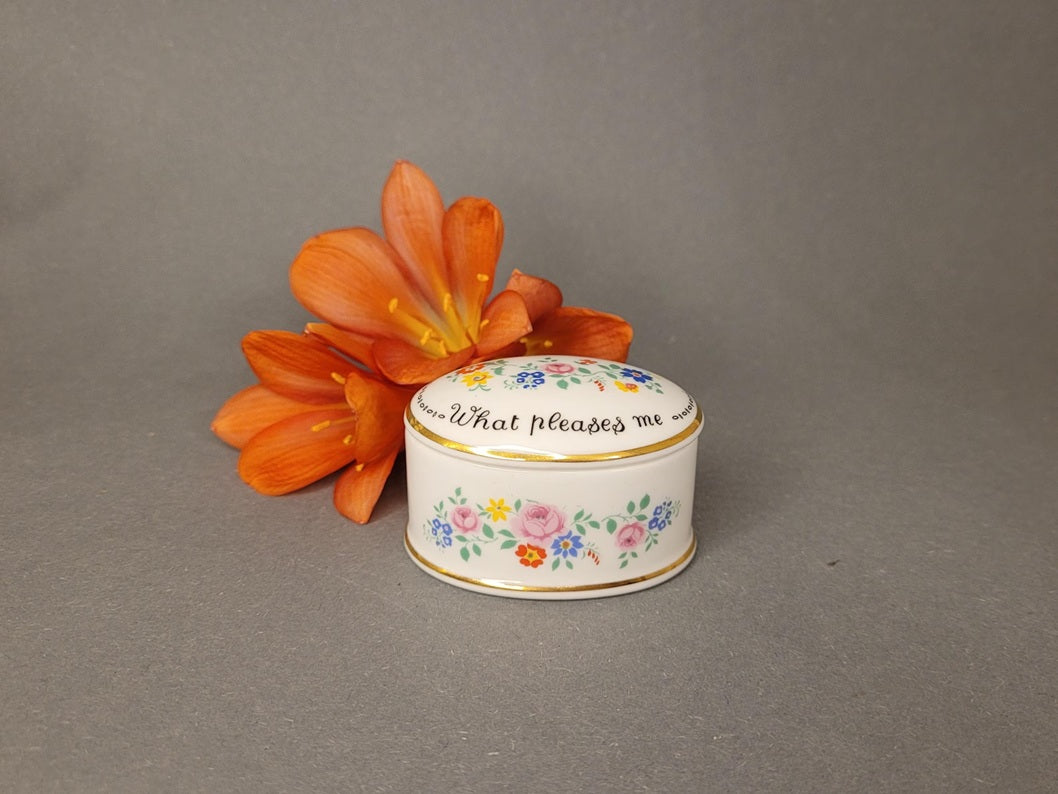 Ovale Pillendose aus weißem Bone China mit bunten Blumen und englischem Sinnspruch neben einer orangen Blume.