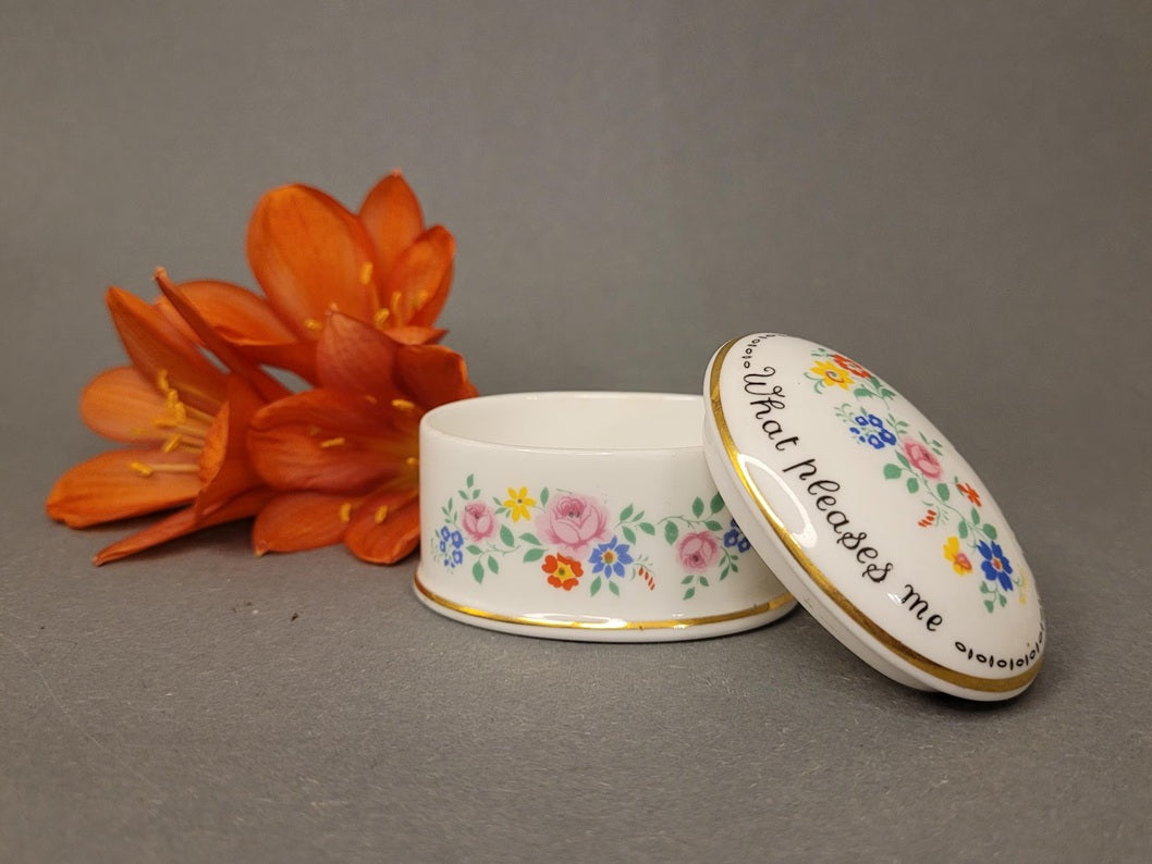 Ovale Pillendose aus weißem Bone China mit bunten Blumen und englischem Sinnspruch neben einer orangen Blume. Geöffnete Seitenansicht.