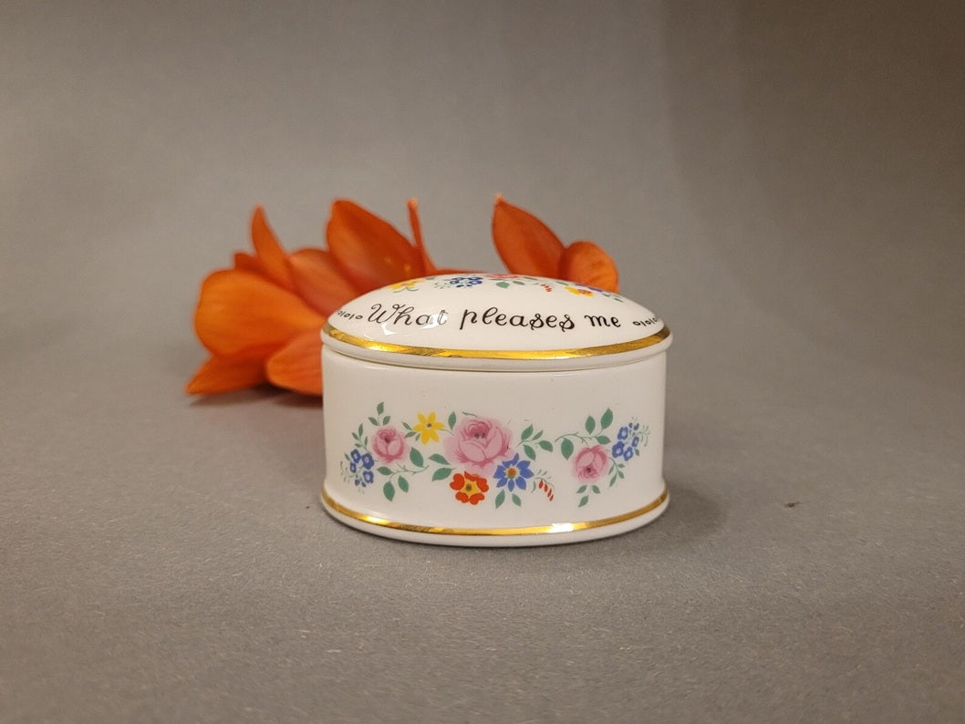 Ovale Pillendose aus weißem Bone China mit bunten Blumen und englischem Sinnspruch neben einer orangen Blume. Seitenansicht.