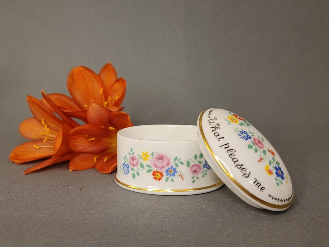 Ovale Pillendose aus weißem Bone China mit bunten Blumen und englischem Sinnspruch neben einer orangen Blume. Geöffnete Ansicht.