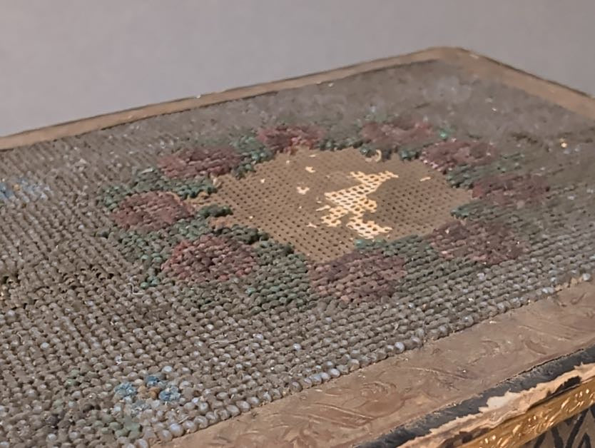 Detail des Deckels: dichter blaugrauer Perlengrund mit Rosenkranz, Mittelfeld mit fehlenden Perlen und freiliegendem Stickkarton