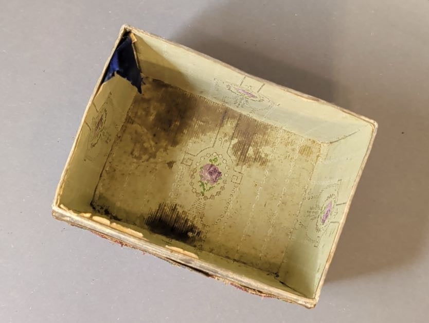 Innenansicht des antiken Schmuckkästchens: hellgrünes Futterpapier mit violettem Blütenmedaillon, dunkle Flecken und eingerissene Ecke