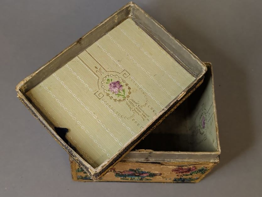 Geöffnetes Perlstickerei-Kästchen: Deckelinnenseite mit mintgrünem Futterpapier und Medaillon mit violetter Blüte, darunter der Korpus