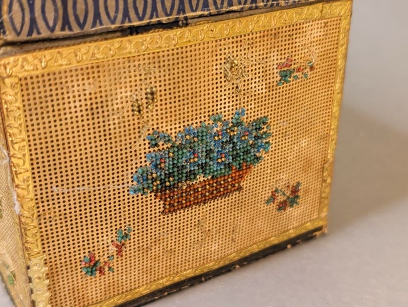 Nahaufnahme Schmalseite mit Perlstickerei-Blumenkorb auf gelochtem Stickkarton, geprägte Goldpapierbordüre und bestoßene Kante