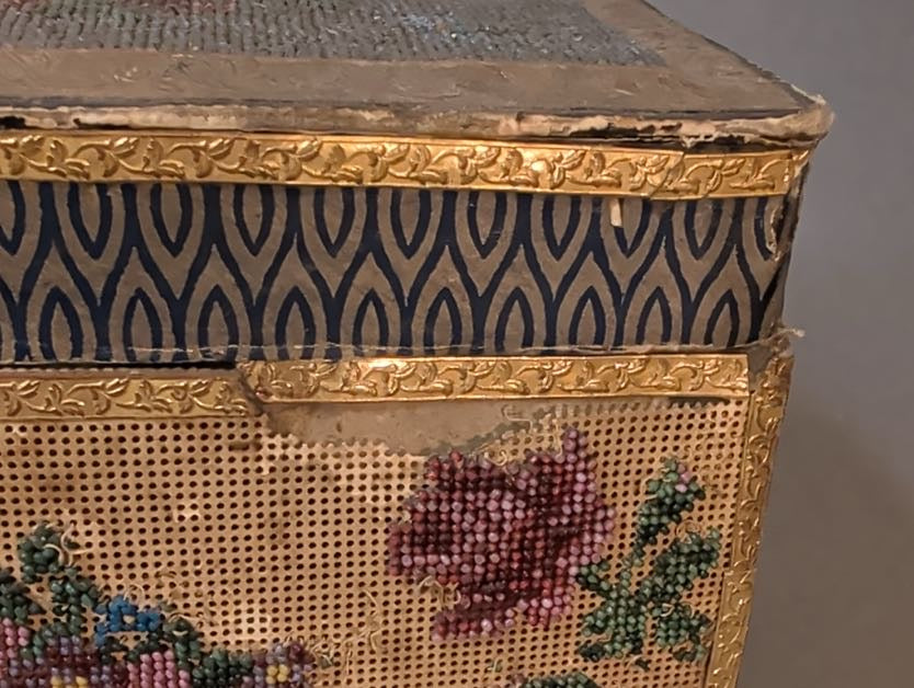 Detail der Deckelecke: bestoßene Papierkante, dunkelblaues Ornamentpapier, geprägte Goldbordüre und rote Rose aus Glasperlen