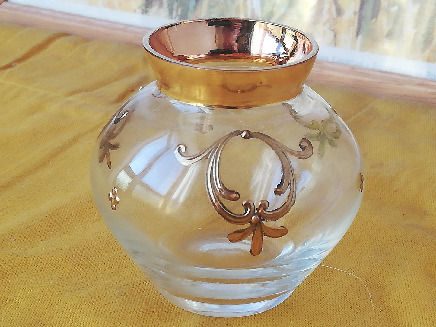 Kleine venezianische Glasvase mit goldenen Reliefs auf einem gelben Tuch vor einem gerahmten Naturbild.