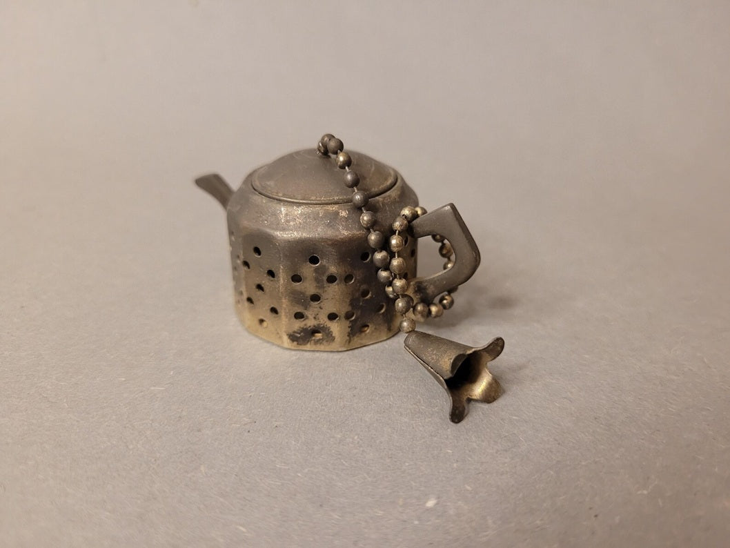 Geschlossenes Miniatur-Teesieb in Teekannenform aus patiniertem Silbermetall auf grauem Grund.