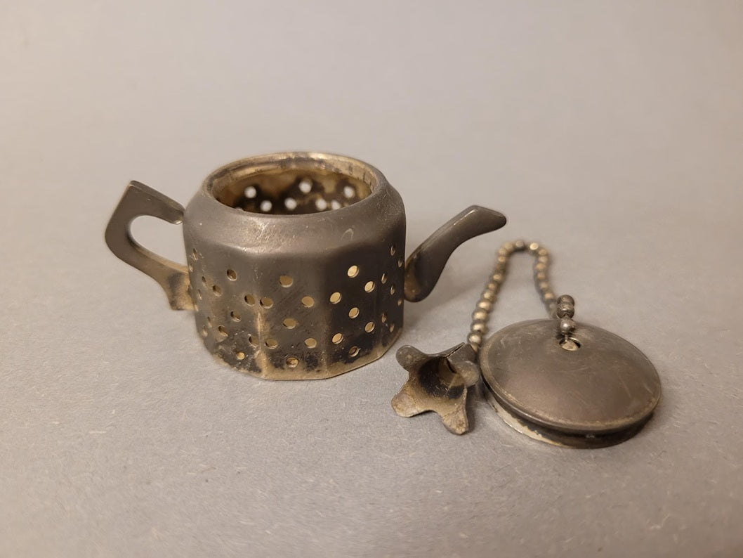 Geöffnetes Teesieb mit abgenommenem Deckel und sichtbarer Kette mit Blumen-Anhänger.